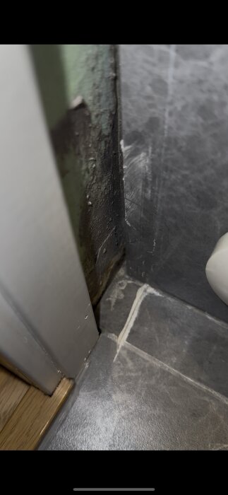 Skadad och fuktig vägg nära badrumströskel med synligt slitet silikon och fog mellan golv och vägg, markerar potentiellt vattenläckage.