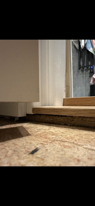 Badrumströskel med borttagen trälist, visar potentiellt fuktskada. Golv och vägg närmare dörrkarmen syns.