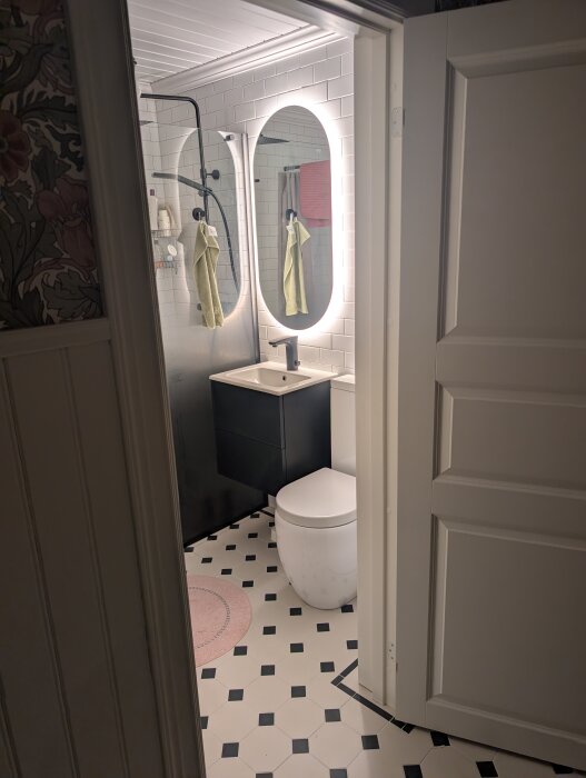 Modernt badrum med vita kakelväggar och svartvit rutigt golv, med ovalt spegel med belysning och en vit toalett.