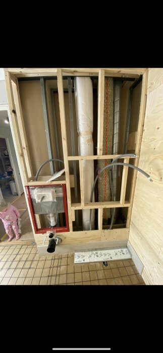 Vägg under konstruktion i våtrum med synliga rör och ramverk, brist på tätskikt kan leda till problem med kakel och silikon/fog. Barn i bakgrunden.