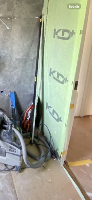 Verktyg och material vid ett byggprojekt med kakel och tätskikt samt en dammsugare och olika verktyg lutande mot väggen.