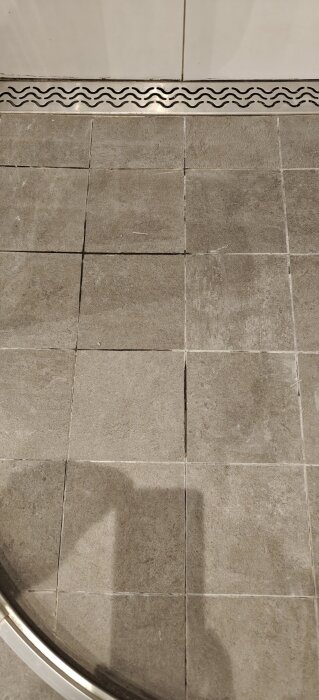 Grå badrumsgolvplattor med fogar, en av dem ser ut att ha släppt något. Synligt duschavlopp med vågformad metallkant.