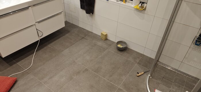 Badrumsgolv med grå kakelplattor, fogar, en svamp, en skål och ett verktyg syns i hörnet av duschen.