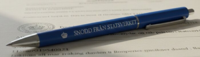 En blå penna med texten "SNODD FRÅN STATSVERKET" ligger på ett papper med text och sigill.