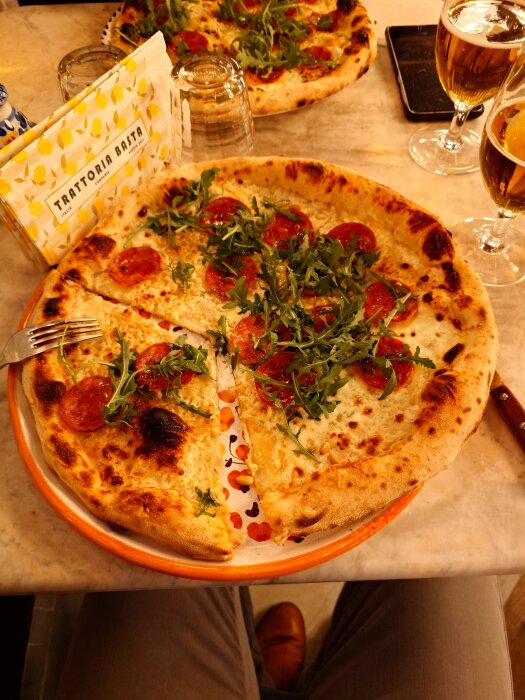 Pizza med pepperoni och ruccola på en tallrik från Trattoria Basta, med två glas öl i bakgrunden.