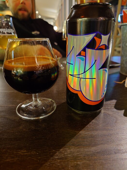 Svart öl i ett glas bredvid en färgglad ölburk på ett bord, med suddig person i bakgrunden.