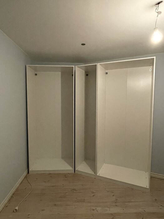 Två vita PAX-garderober mot blå vägg i ett hörn, med mellanrum mellan garderoberna. Lamphållare i taket och sladd på golvet.