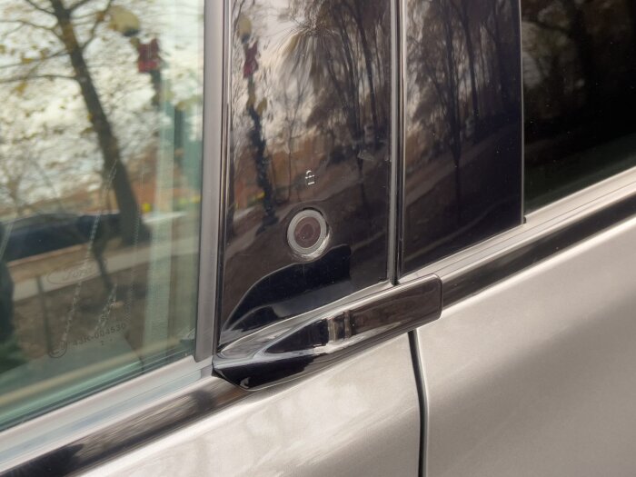 Närbild på dörrknapp för en elbil, troligen en Ford Mach-E, med reflektioner av träd och lyktstolpar på fönstret.