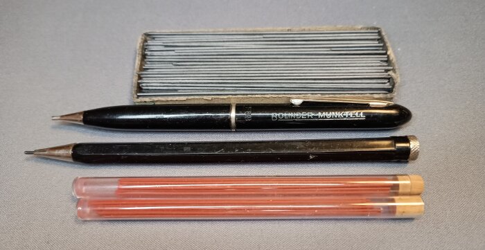 Gammaldags blyertspennor och reservstift i glasrör, märkt "BOLINDER MUNKTELL", med stiftlåda vid sidan på en duk.