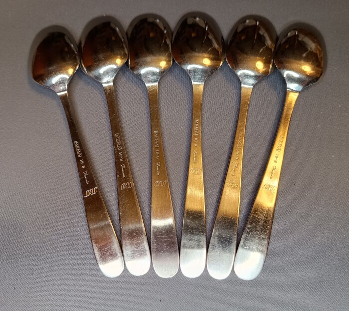 Sex metallskedar med ingraverad text, placerade sida vid sida på en grå yta.