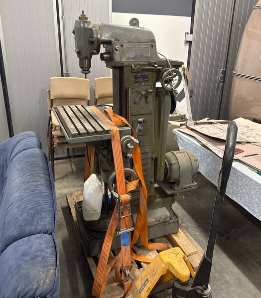 Metallfräs Deckel FP1, stående i ett garage bredvid en soffa. Maskinen är säkrad med orangea spännband och står på en pall.