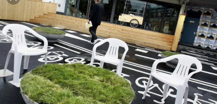 Tre vita plaststolar placerade på en cykelväg med grönska. En person går förbi i bakgrunden vid en byggnad.