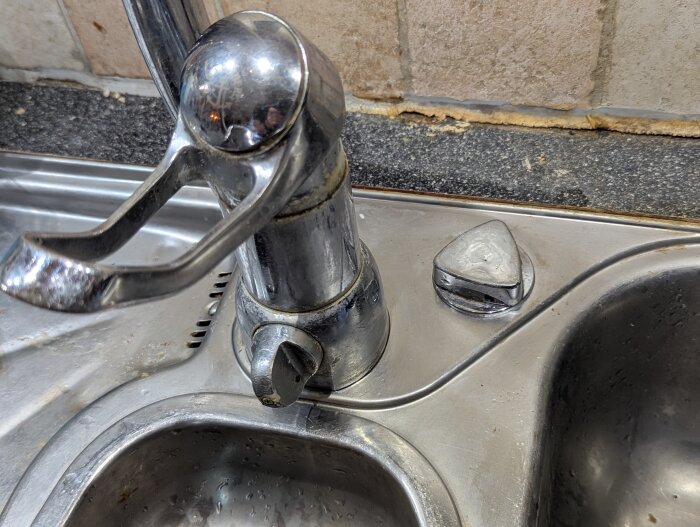 Kran med trasigt vred vid diskhon med synlig bottenventil och köksbänk i metall.