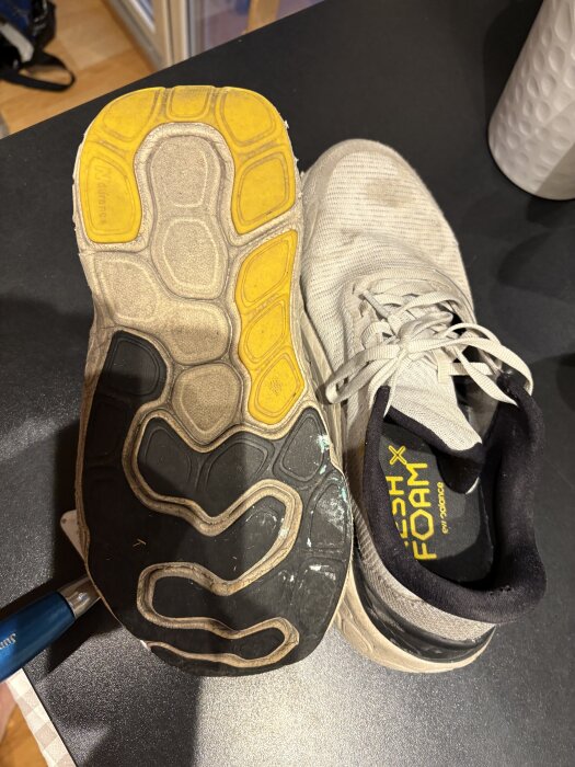 Ett par vita löparskor på en mörk yta, med en sula uppåt som visar mönster i gult och svart samt texten "Fresh Foam" inuti skon.