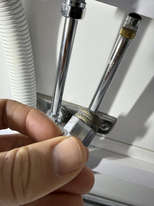 Hand håller en metallkoppling med gängor täckta av grön pasta, bredvid två anslutna metallrör i ett VVS-system.