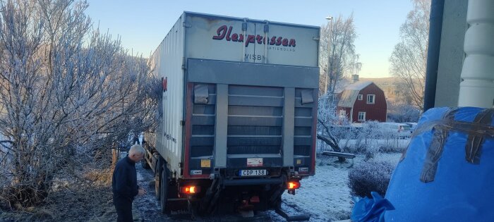 Lastbil står parkerad på vinterväg framför ett rött hus; en person står bredvid. Träden och marken är frosttäckta.