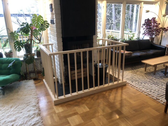 Öppen spis i vardagsrum med trägrind som skydd. Eldstad omgiven av soffor och växter. Parkettgolv och stora fönster i bakgrunden.