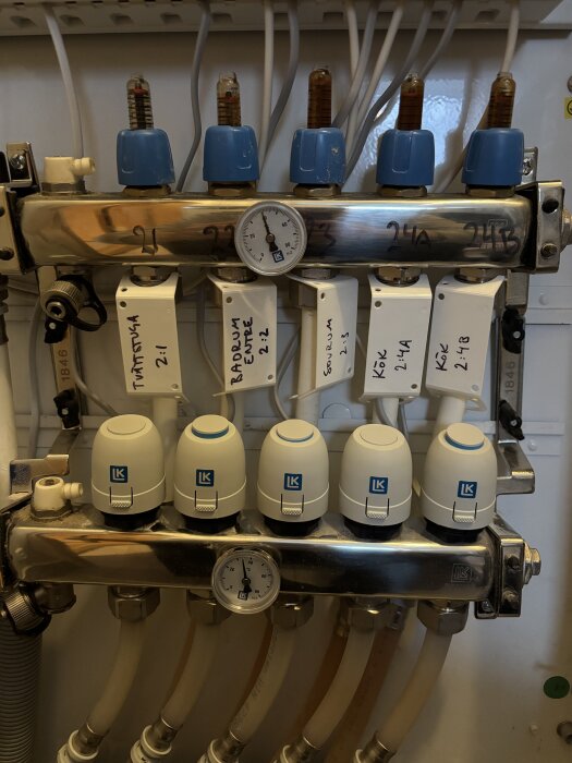 Bild av ett golvvärmesystem med LK-termostater och ställdon. Det finns en tryckmätare och etiketter för olika rum, inklusive kök och badrum.