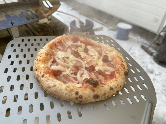 En hemmagjord pizza med skinka, lök och korv på ett pizzagaller utomhus i vintrig miljö.