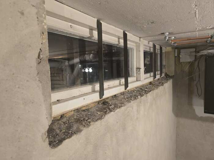 Gammalt fönster med uttorkad träram och murbruksskador, träramen är krackelerad och muren har grov textur bland vindlande rör ovanför.