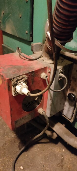 Röd pelletspanna med kablar, kontakt och ventilationshål, kopplad till en grön värmepanna i ett pannrum.