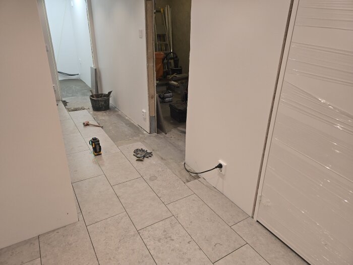 Renoverad korridor med kakelgolv, handskar på golvet, hink och verktyg synliga, spacklingsarbeten pågår i bakgrunden.