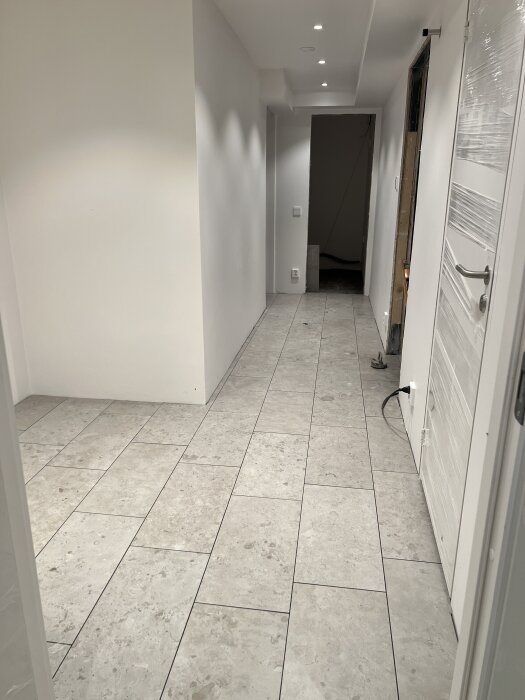 Korridor med nyplattsatt golv i ljus sten, vita väggar och dörröppning i bakgrunden.