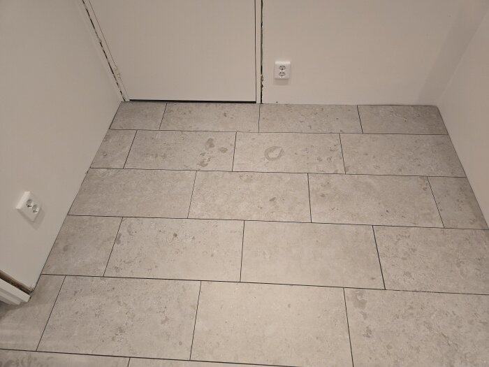 Grått klinkergolv i en korridor med vita väggar och eluttag.