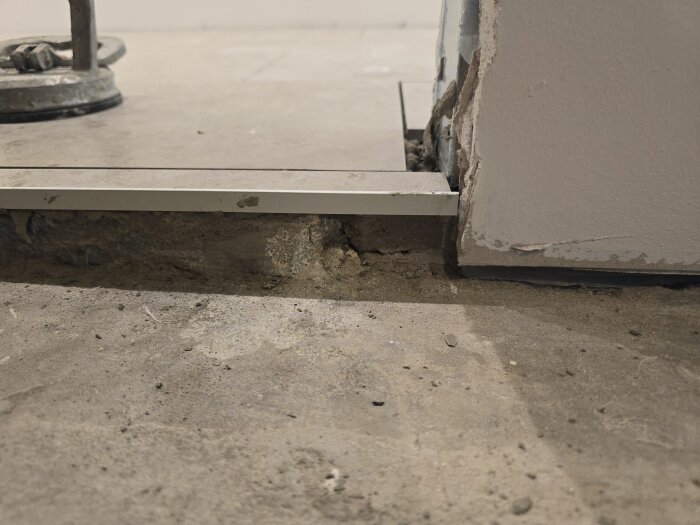 Golvrand i korridoren med synlig nivåskillnad mellan rum, mindre skada på väggen och golvets yta, redo för slutlig justering och plattsättning.