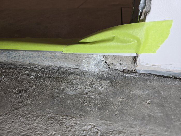 Närbild av golv med tejp runt en dörrtröskel, visar ojämnheter och sprickor i betong. Förberett för ytterligare flytspackling och arbete.