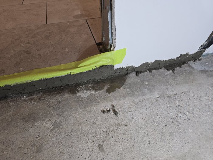 Golv med nylagd lagningsbruk vid en tröskel i en korridor, gult tejp skyddar väggen.