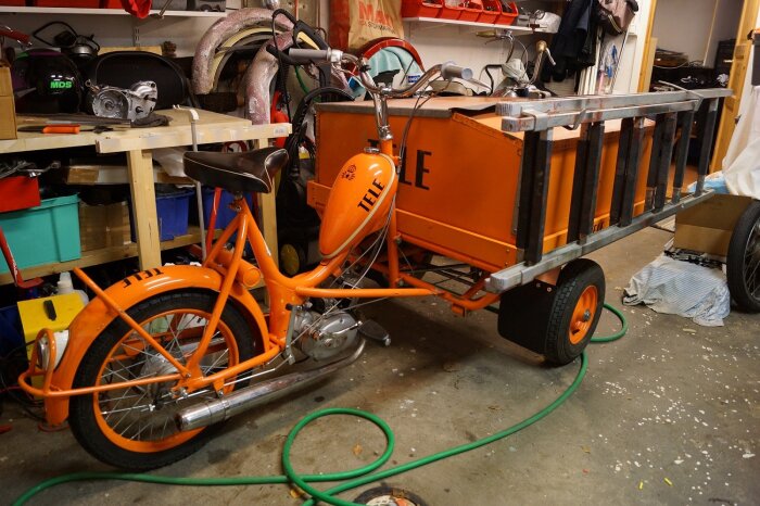 Orange flakmoped från 60-talet med texten "TELE", stående i ett garage med diverse verktyg och delar runtomkring.