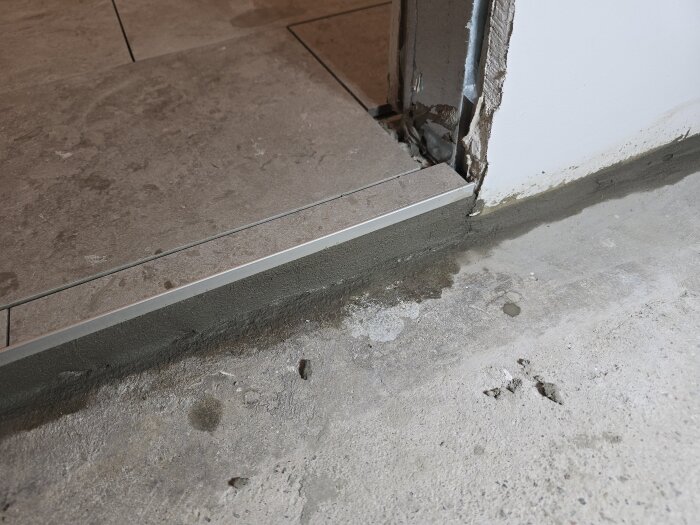 Tröskel av metall mot kaklat golv, med synlig bruk eller fogning på betonggolv. Väggen är delvis ofärdig vid dörrkarmen.