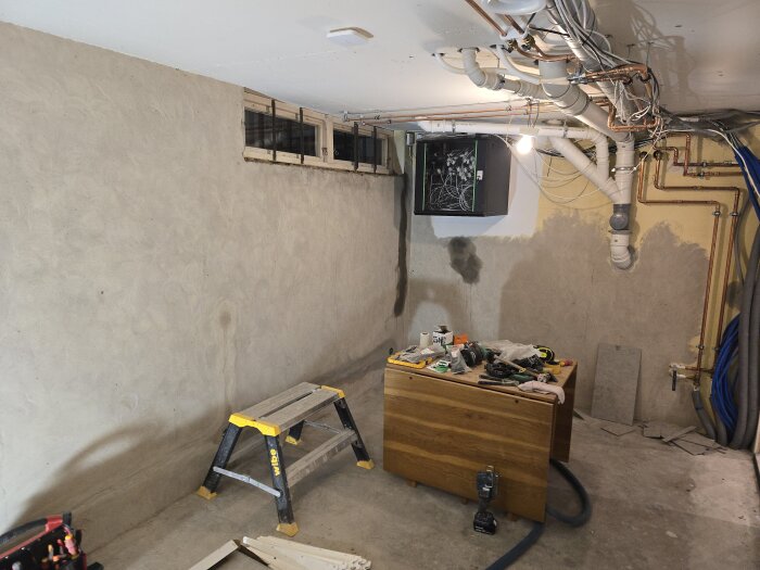 Källarutrymme under renovering med olika verktyg på en träbänk, ett litet fönster och synlig rördragning i taket.