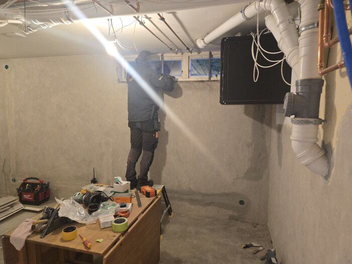 Person arbetar på fönsterinstallation i källarutrymme med verktyg och rör dragna längs taket. Verktygslåda och material utspridda på bord.