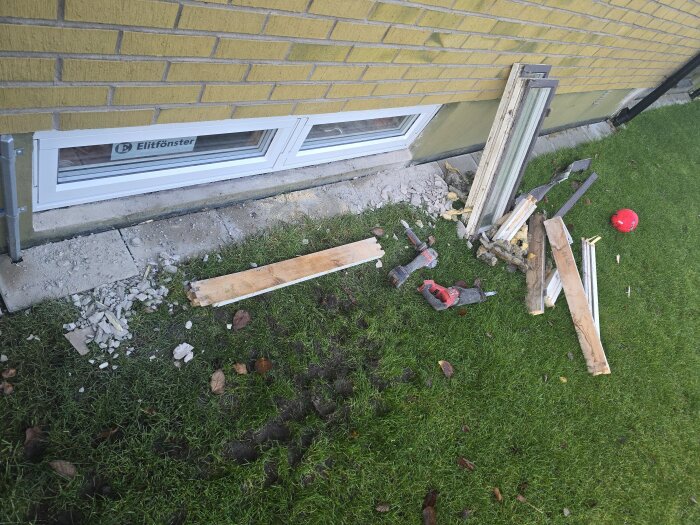 Byggmaterial och verktyg ligger framför ett nyinstallerat vitt fönster på en gräsmatta. En röd hjälm syns till höger och en murad vägg i bakgrunden.