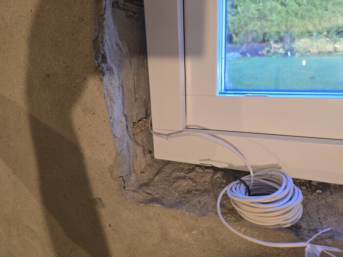 Nytt fönster installerat i en källarvägg; vit kabelrulle bredvid ramen, ansluten för magnetkontakt. Fasaden är putsliknande och landskap visas utanför.