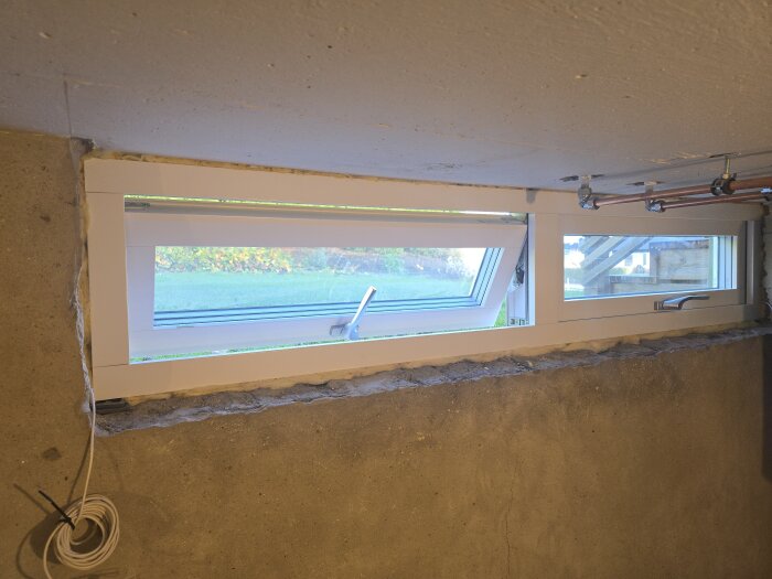 Nytt vitt fönster installerat i en källarvägg, med synliga rör och kabel på väggen. Fönstret har två öppningar och en modern design.