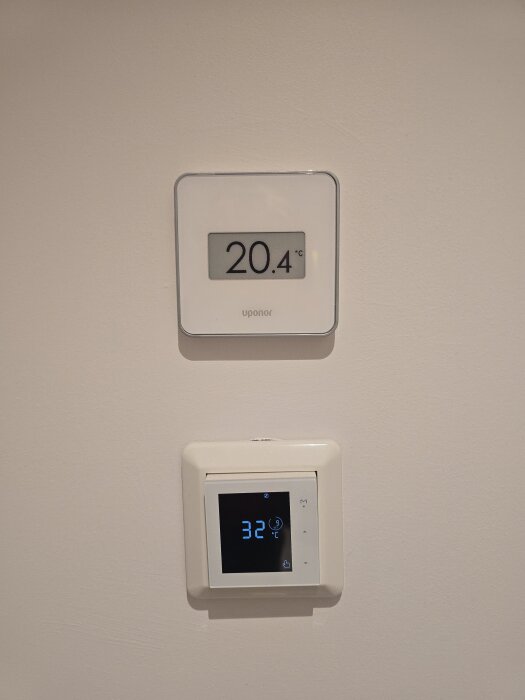 Två termostatpaneler på väggen, den ena visar 20,4°C och den andra 32°C, indikerar olika temperaturer för rummet och golvvärmen.