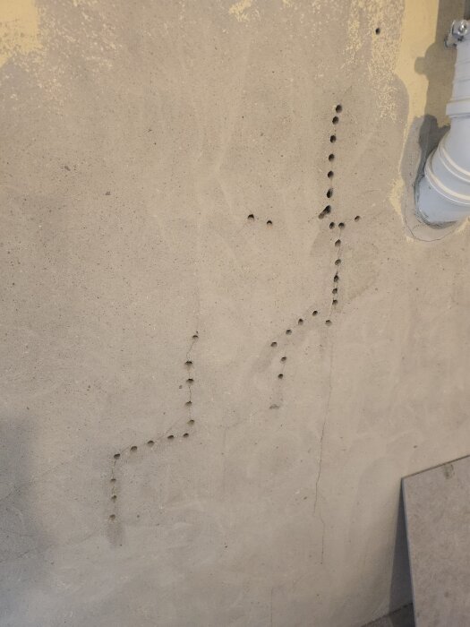 Vägg med borrhål i ett mönster bredvid en vit rörledning i ett rum under renovering.