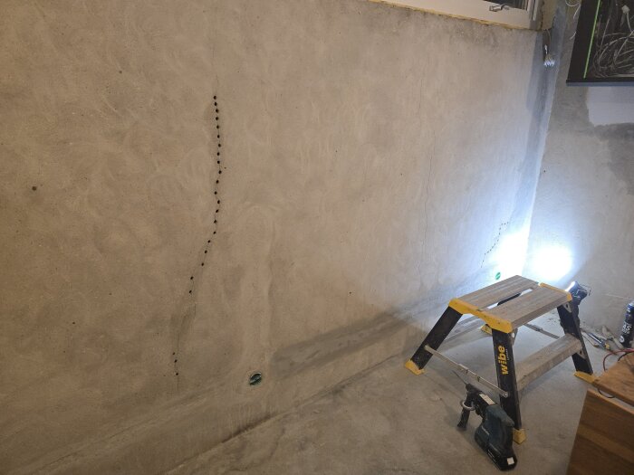 Betongvägg med sprickor bredvid en arbetsstege och en borrmaskin på betonggolvet, belyst av en arbetslampa.