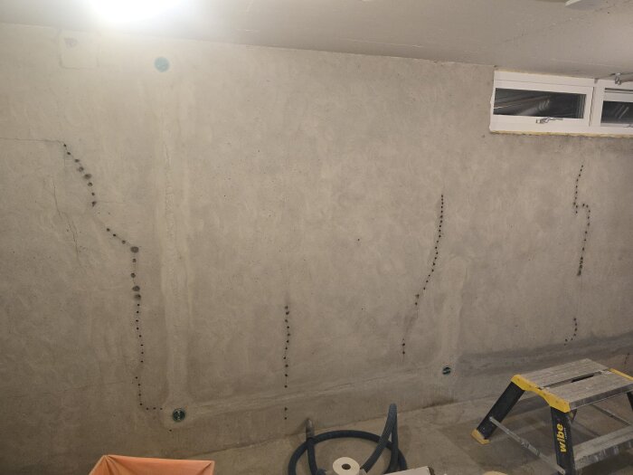 En betongvägg med sprickor och borrade hål markerad med murbruk, intill en stege och verktyg. En ventilationsöppning syns uppe till höger.