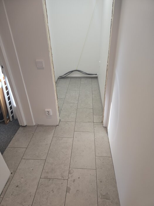 Korridor med nytt klinkergolv och vitmålade väggar. Golvet har längsgående fogar och ansluter till två dörröppningar. Kabel löper längs väggen.