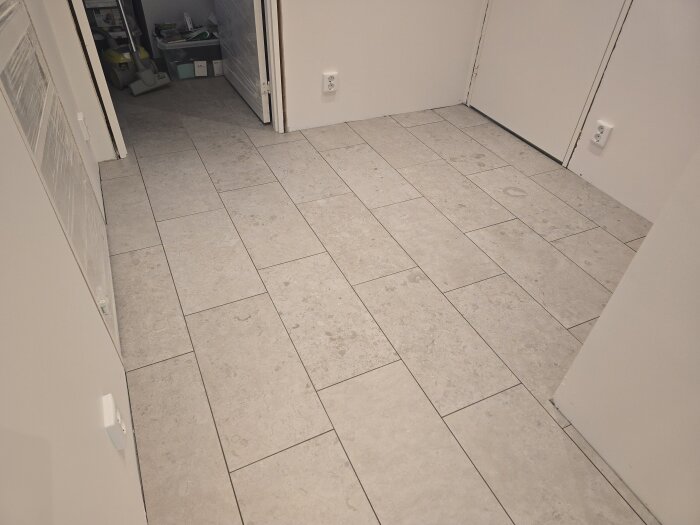 Nyförlagda ljusa kakelplattor på ett källargolv med en korridor och rum i bakgrunden; eluttag syns på väggen.