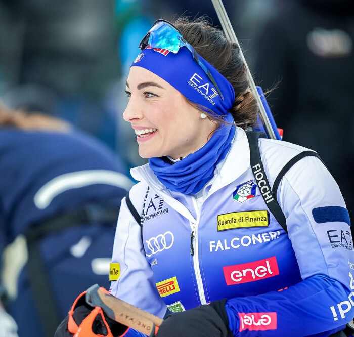 Kvinna i blå skiddräkt och solglasögon i pannan ler, med sportutrustning i bakgrunden.