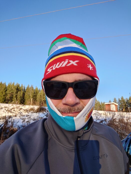 Person med mössa och solglasögon åker skidor i solig vintermiljö, med snö, trädsiluetter och klarblå himmel i bakgrunden.