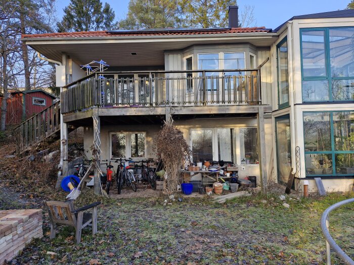 Under balkongen på ett hus förvaras cyklar och diverse föremål oskyddade från väder, omgärdat av höstlöv och gräs.