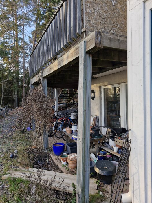 Under balkongen förvaras olika föremål inklusive krukor och cyklar, oskyddade mot regn, med en trästruktur och natur i bakgrunden.