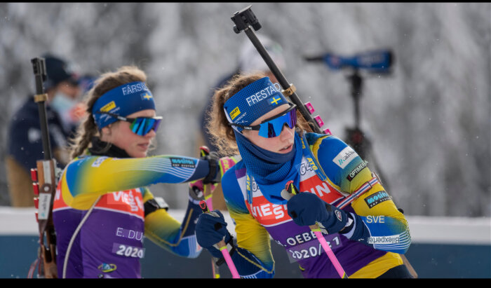 Två kvinnliga skidskyttar med gevär på ryggen, klädda i svenska landslagsdräkter, är i rörelse under en tävling.