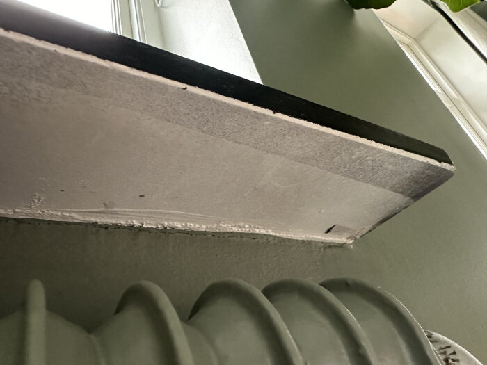 Undersidan av en hylla med svart ytskikt, där det inre materialet är synligt och visar annan färg än det yttre lagret, intill ett fönster och ett element.